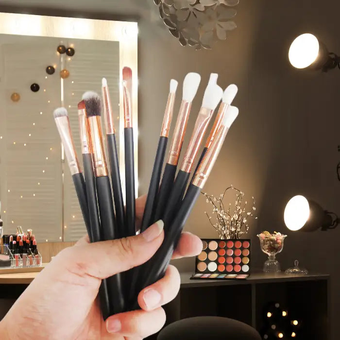 Professional makeup brushes with black handles and rose gold ferrules held at a makeup station with palette, vanity lighting and decorative elements.