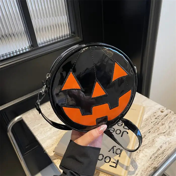 Glossy black Pumpkin Head Purse with orange pumpkin face held in hand near tabletop decor.