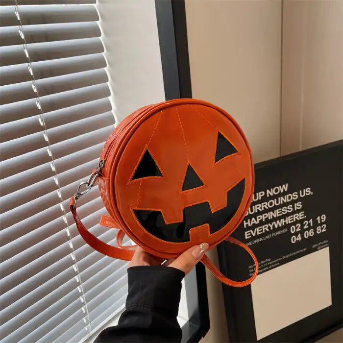 Shiny orange Pumpkin Head Purse with black face detailing, held in hand near blinds and frame.