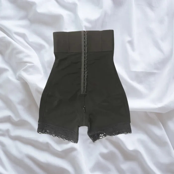 Full front view of black shapewear with high waistband and lace leg trim on white bedding.