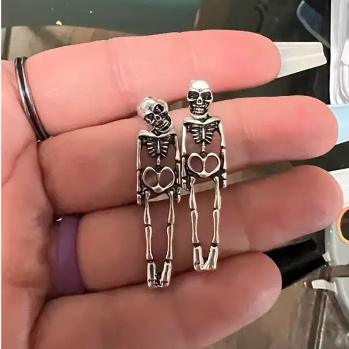 Close-up of silver skeleton earrings shown in hand, featuring detailed bone structure and skull top.