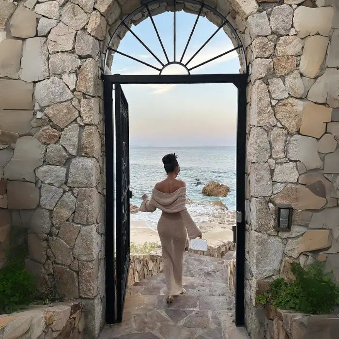 Women's one-shoulder long-sleeve knitted vacation dress in beige walking through stone gate to ocean.