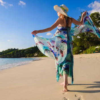 Summer dresses with watercolor pattern flowing elegantly as woman walks barefoot on tropical beach shore wearing wide-brimmed sun hat