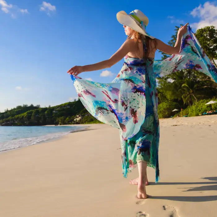 Summer dresses with watercolor pattern flowing elegantly as woman walks barefoot on tropical beach shore wearing wide-brimmed sun hat