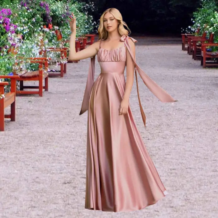 Blonde model poses elegantly in a blush pink satin dress. This long gown features tie shoulders and a flattering empire waist.