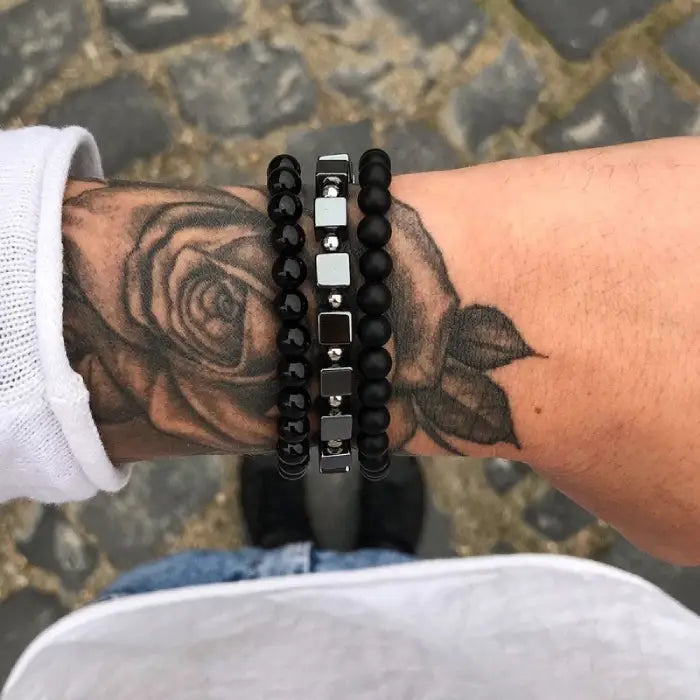 mens bracelet stack.  Person wearing two bracelets with black beads and silver square accents on a stone pavement background.