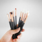 Professional makeup brushes featuring black handles and copper ferrules held against neutral background, showcasing various shapes for makeup application.