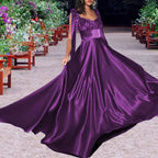 Woman showcasing an elegant purple satin dress in a garden setting. The formal gown has tie straps and a flowing maxi skirt.
