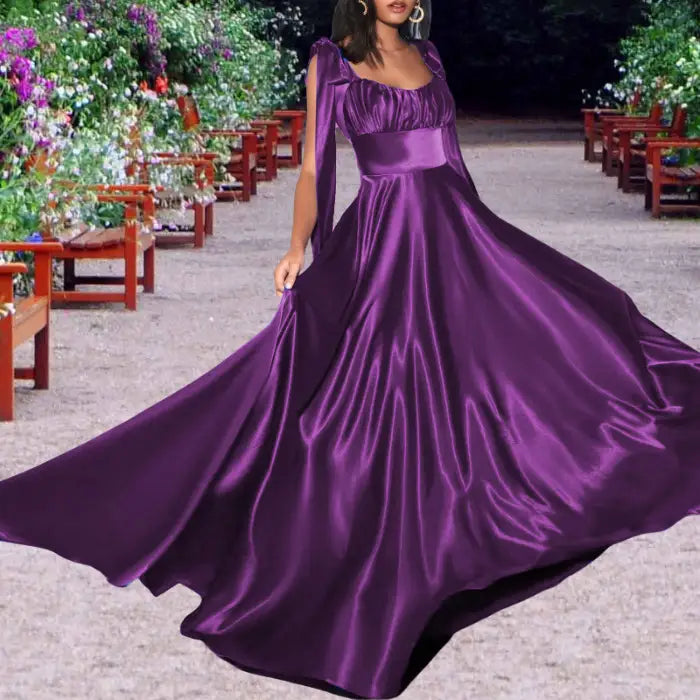 Woman showcasing an elegant purple satin dress in a garden setting. The formal gown has tie straps and a flowing maxi skirt.