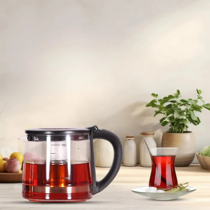 Glass teapot filled with tea on a wooden table next to a glass of tea and a potted plant.