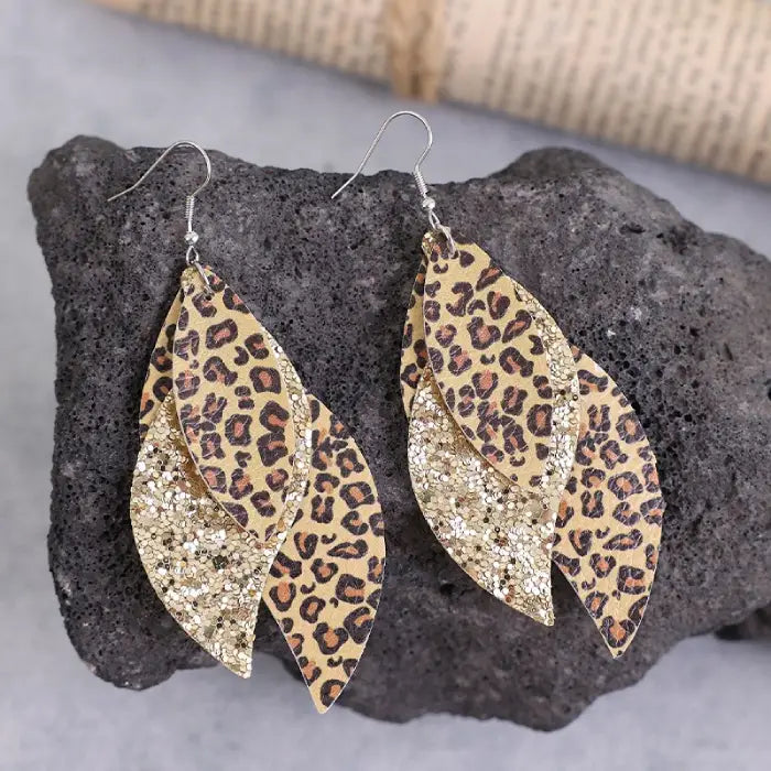 A pair of layered leaf-shaped earrings with leopard print and glitter accents displayed on a gray textured background.