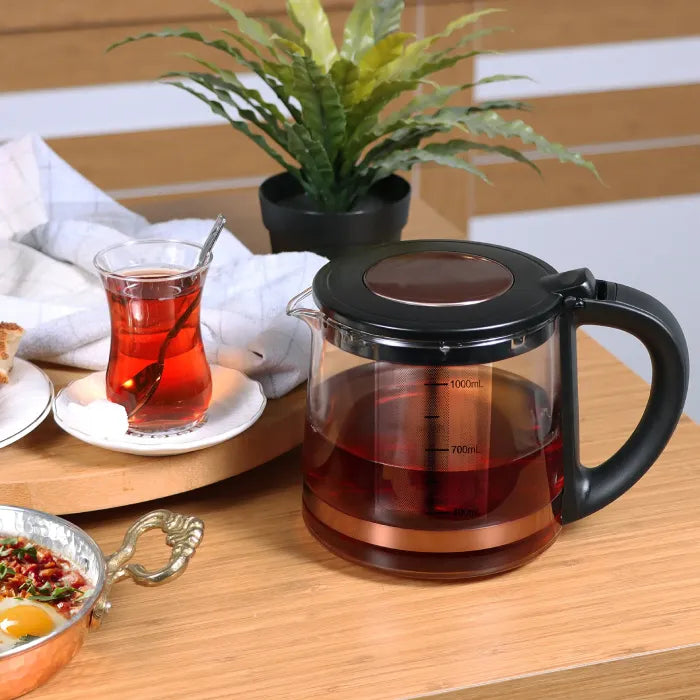 Electric tea maker with glass chamber and rose gold accents serving red tea alongside breakfast items and plant.