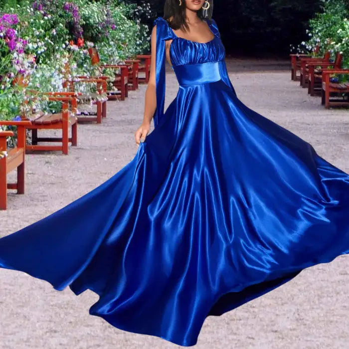 Model twirls in a vibrant royal blue satin dress outdoors. This long formal gown features tie shoulders and a full skirt.