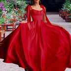 Model displays a vibrant red satin dress with full skirt outdoors. Features romantic tie straps, perfect formal evening wear.