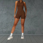 activewear jumpsuit in chocolate brown with front zipper closure and mock neck design styled with white sneakers against gray wall
