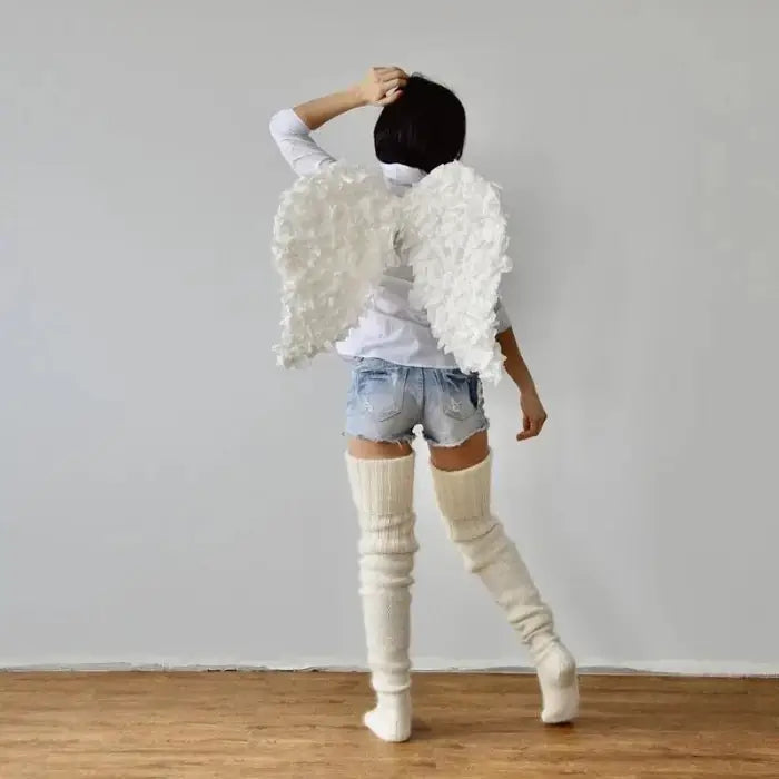 Person wearing white feathered angel wings, denim shorts, white knee-high socks, standing on wooden floor