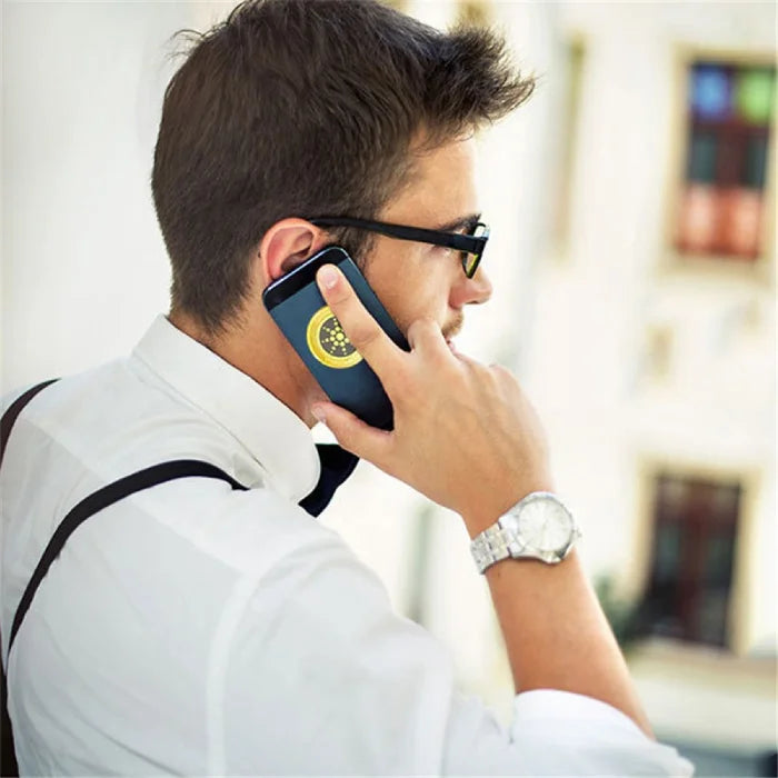Anti radiation sticker on phone held to ear by man in white shirt and sunglasses outdoors.