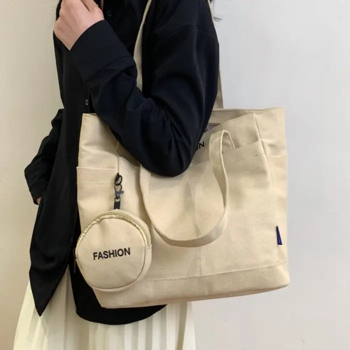 Beige canvas tote bag with mini pouch labeled "FASHION," carried by a woman in a black shirt.