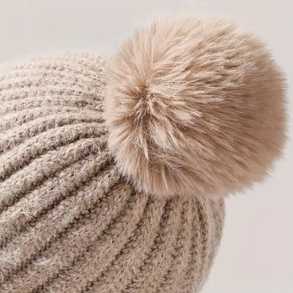 Side view of a beige fuzzy knit winter hat with a large fur pom-pom on top 