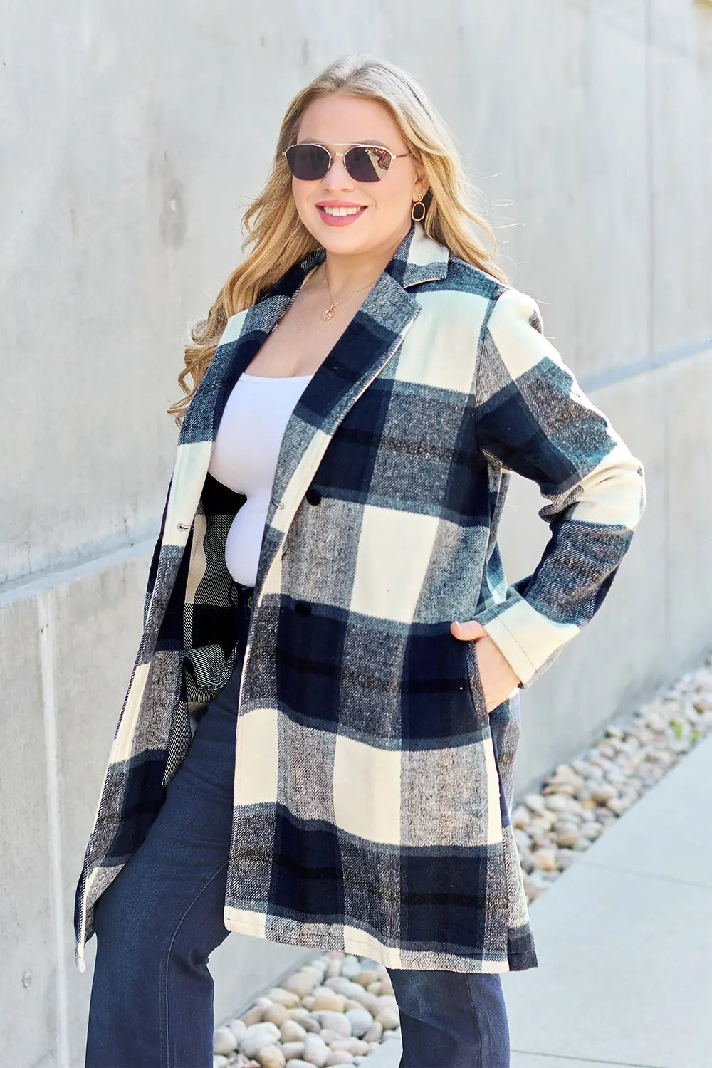 Woman wearing a black and white plaid wool coat styled with flared jeans and a white top, smiling outdoors near a concrete wall.