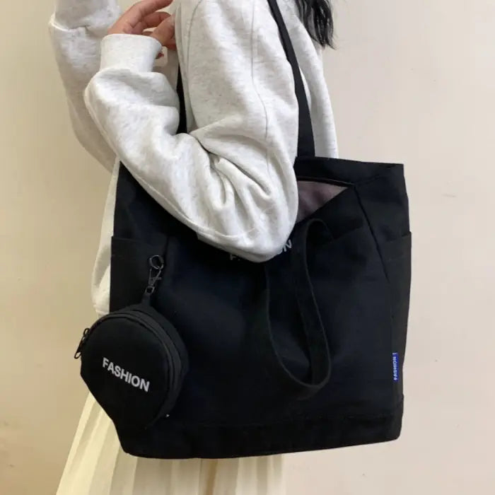 Black canvas tote bag with mini pouch labeled "FASHION," carried by a woman wearing a gray sweatshirt.