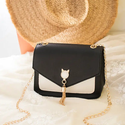 Black crossbody bag with gold chain, cat charm, and tassel detail placed on lace fabric with a straw hat background.