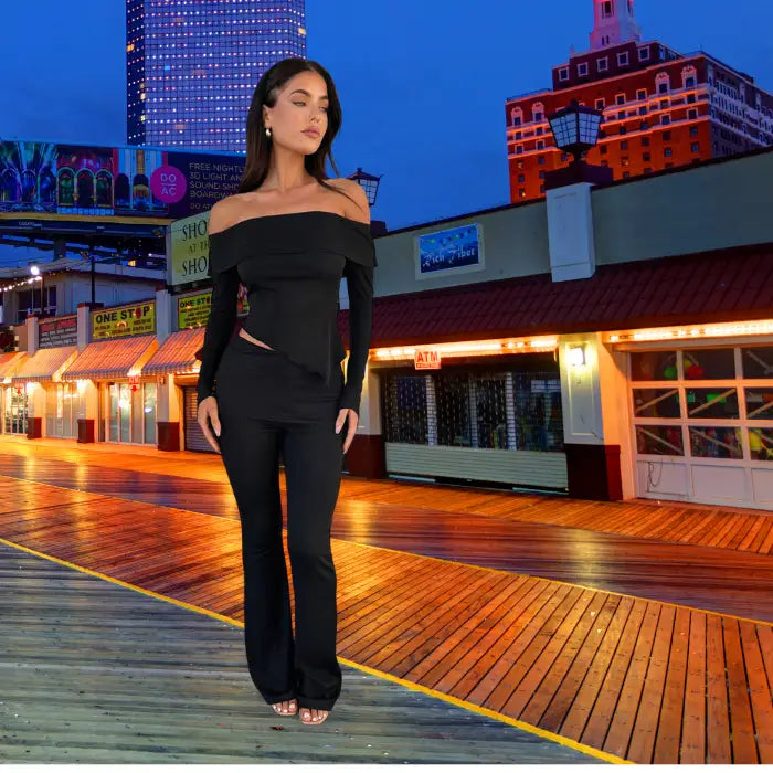 Black formal outfit with off-shoulder design illuminated by city lights on wooden boardwalk against vibrant nighttime skyline backdrop