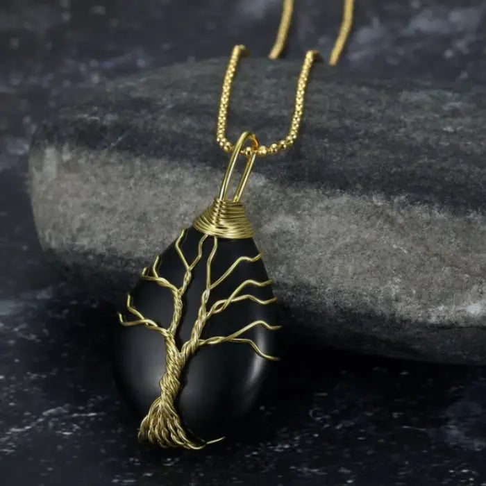 Black teardrop gemstone pendant with gold wire tree of life design on gold chain and stone background.