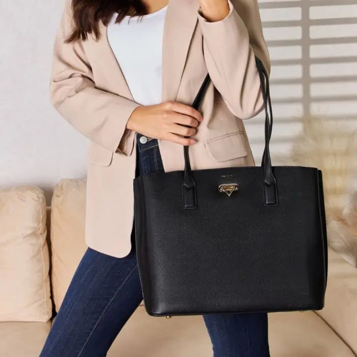 Close-up of a black leather tote bag with gold emblem, held by a person wearing a beige blazer and jeans