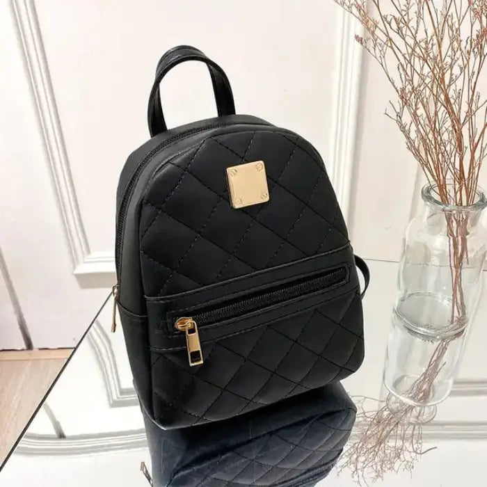 Black quilted mini backpack with gold metal plate and front zipper pocket, displayed on reflective surface next to glass vase