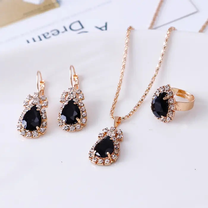 Black teardrop gemstone jewelry set with necklace, earrings, and ring in gold surrounded by crystals.