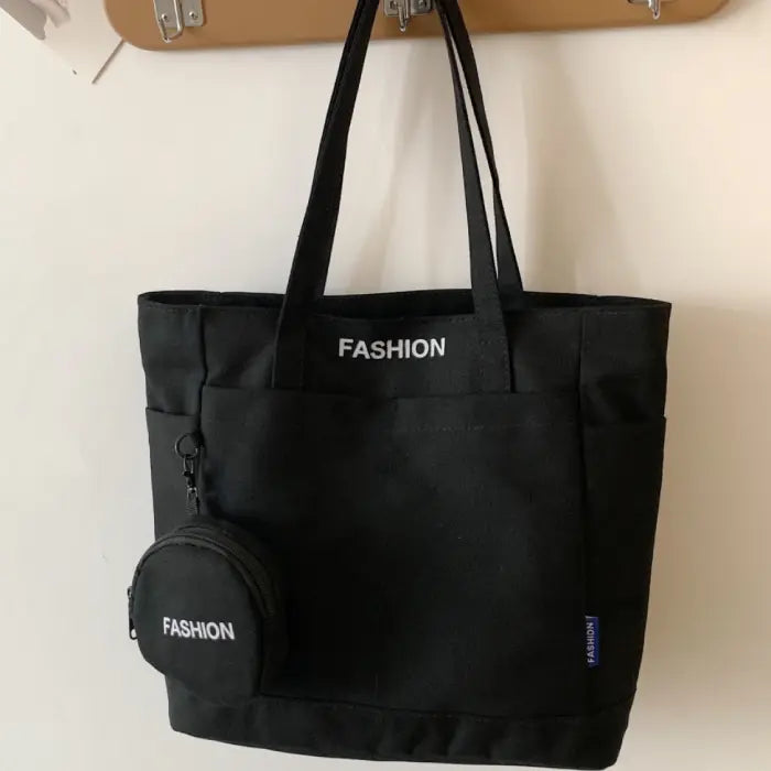 Sleek black tote bag with mini pouch labeled "Fashion," hanging on a modern wooden wall hook.