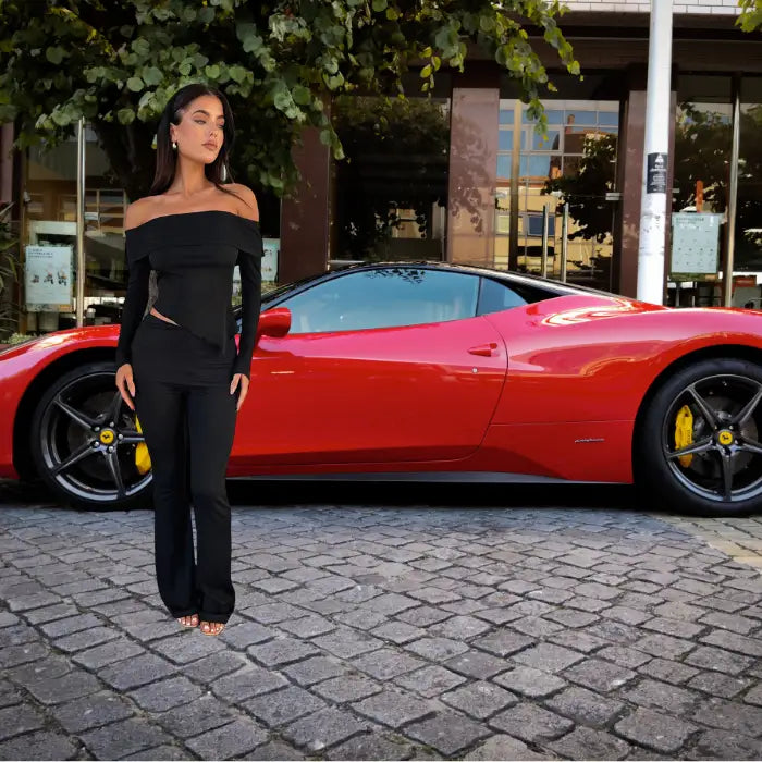 Black two piece outfit featuring off-shoulder top with side cutout and flared pants shown with luxury red sports car on cobblestone street
