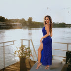 Blue gown with sparkly sequins in royal blue featuring high thigh slit, worn by model posing at riverside sunset setting.