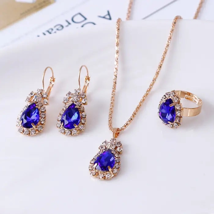 Blue teardrop gemstone jewelry set with necklace, earrings, and ring, accented with gold and crystals.