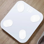 White Bluetooth body fat scale with four round sensors showing weight on LED display placed on wood floor for home fitness use
