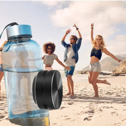  bluetooth speaker shown with water bottle on beach while people enjoy outdoor activities in background