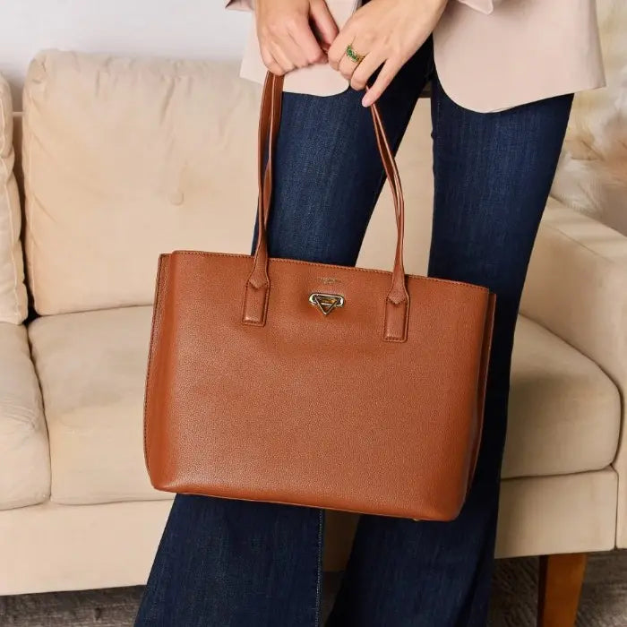 Woman holding brown leather tote bag while standing near beige sofa, wearing beige coat and blue jeans