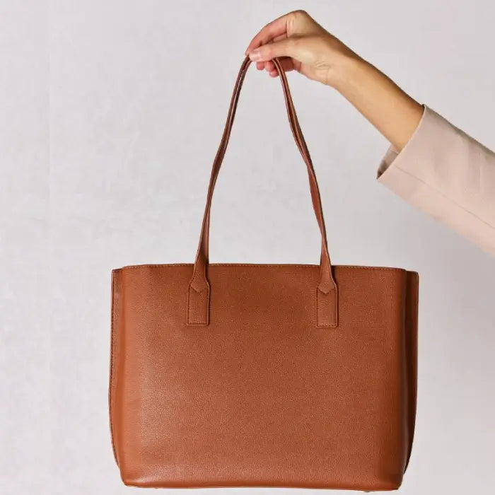 Brown leather tote bag held by hand against white background, displaying its simple yet elegant design