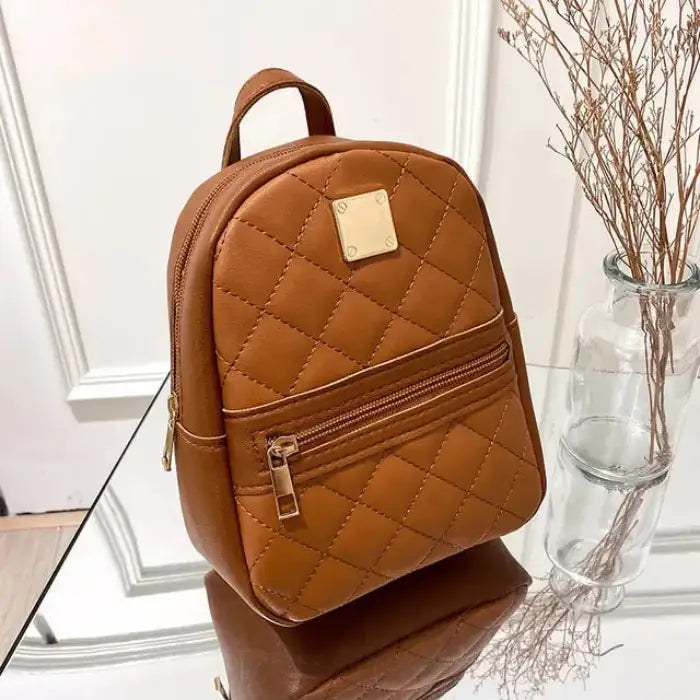 Tan quilted mini backpack with gold metal plate and front zipper pocket, displayed next to glass vase with dry branches