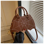 Brown leather handbag with white spiderweb stitching, bat-wing shape, held by hand with red nails near white blinds