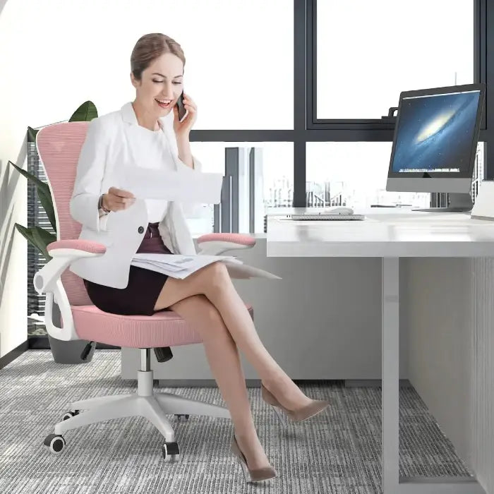 Professional woman in white blazer sitting on pink office chair, working on computer and talking on phone in bright office