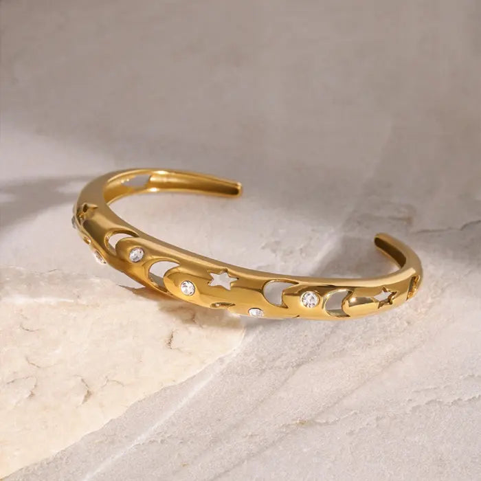 Celestial-themed gold plated bangle with moon and star cutouts, adorned with stones, placed on a marble surface.