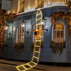 Christmas ladder lights with Santa climbing building front decorated with garlands and warm lighting.