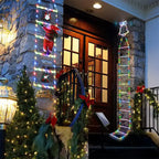 Christmas ladder lights with Santa climbing near stone porch decorated with garland and bows.