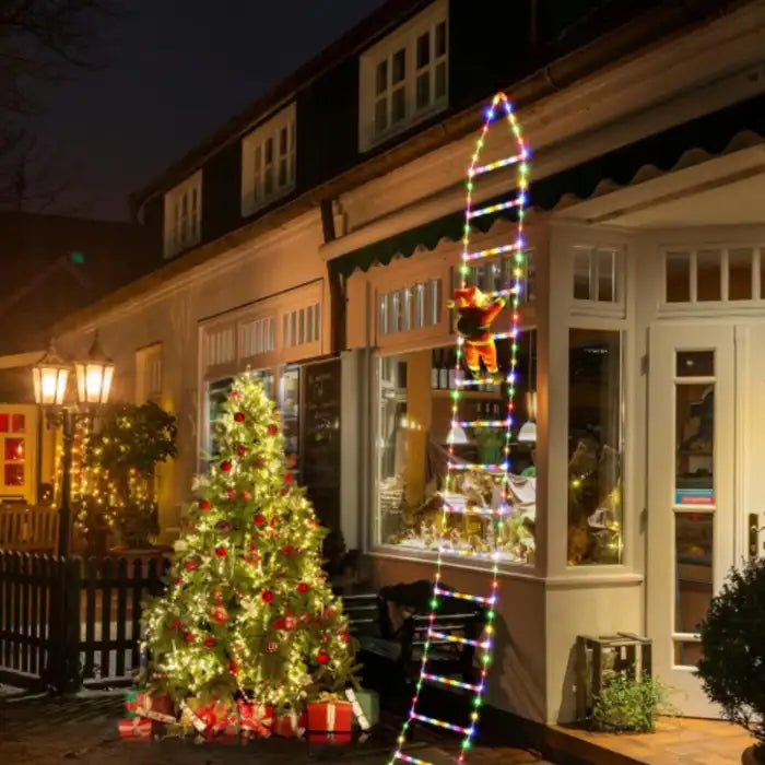 Christmas ladder lights with Santa climbing storefront window next to decorated tree and gifts.