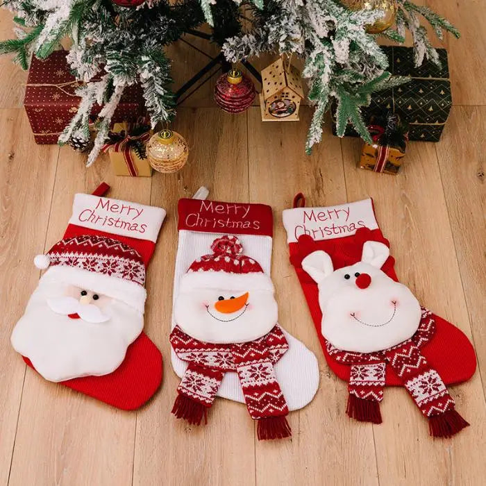 Three Christmas stockings with Santa, snowman, and reindeer designs laid under decorated Christmas tree with wrapped presents