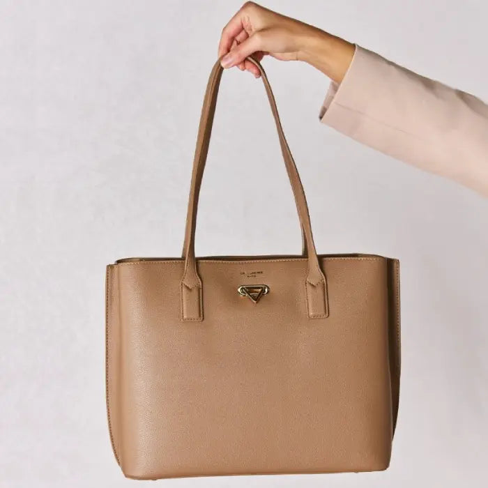 Tan leather tote bag with gold hardware held by a hand in a beige sleeve, showcasing the bag's structure and design