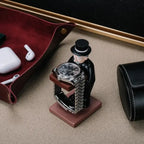 collector watch display figurine holding a watch, surrounded by desk accessories like AirPods and a leather tray.