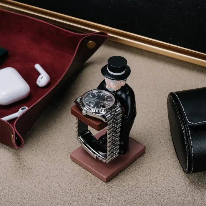 collector watch display figurine holding a watch, surrounded by desk accessories like AirPods and a leather tray.